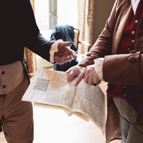 Two unidentified men in colonial dress pointing at and gesturing over a newspaper