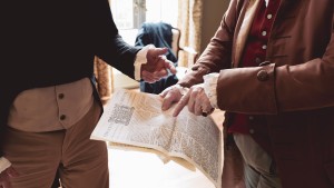 The hands of two men dressed in colonial clothing gesture and point at a colonial newspaper