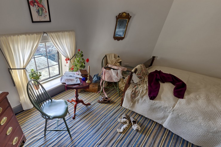 A bedroom with a striped carpet, a low window, a chair, and a small table crowded with books and papers