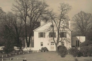 A large two-story, white brick house with a large classical pediment, several chimneys, and an extensive lawn in which a black poodle is laying.