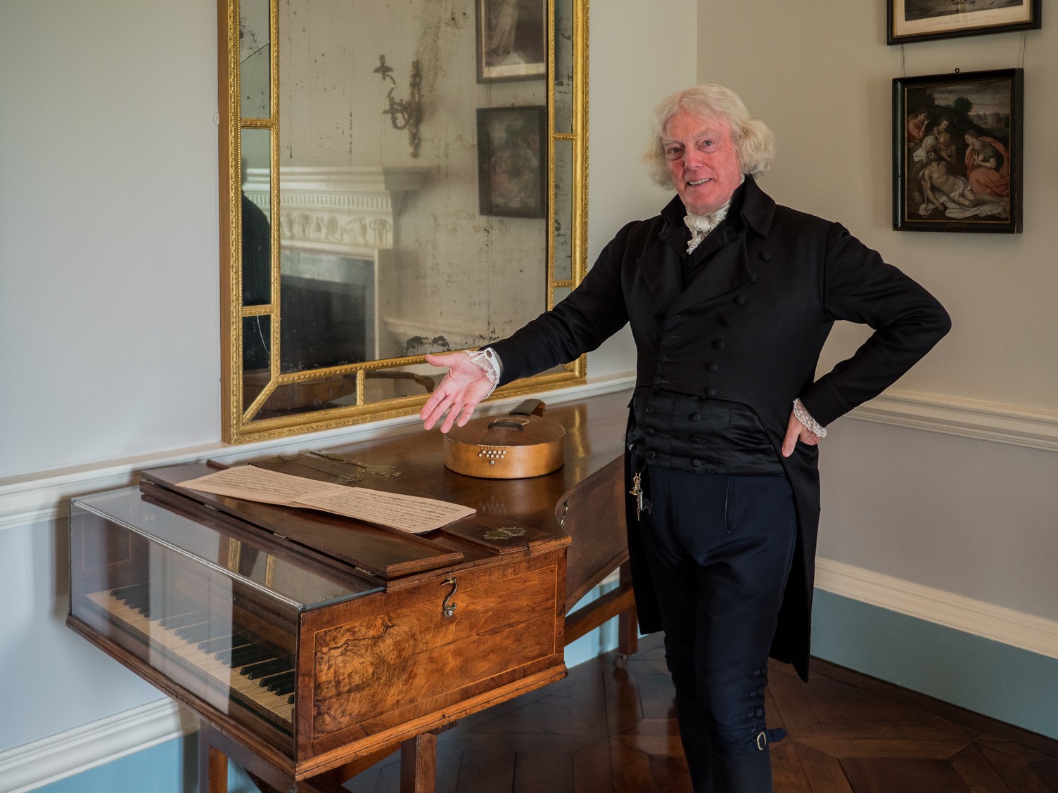 Bill Barker portraying Thomas Jefferson, dressed in a 19th century black frock coat and breeches gesturing towards an antique harpsichord